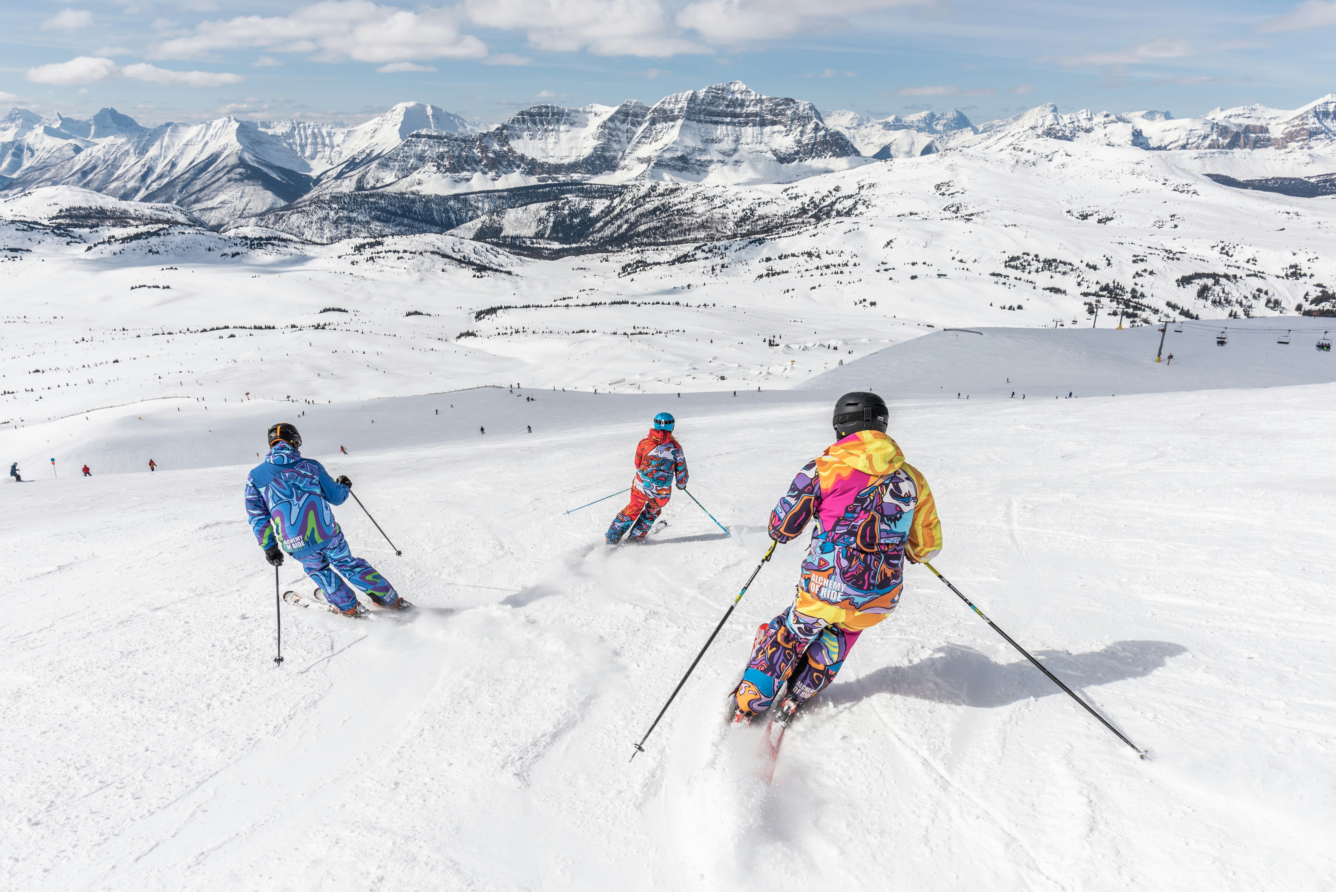 Tres esquiadores bajan juntos una ladera abierta frente a un mar de cumbres nevadas