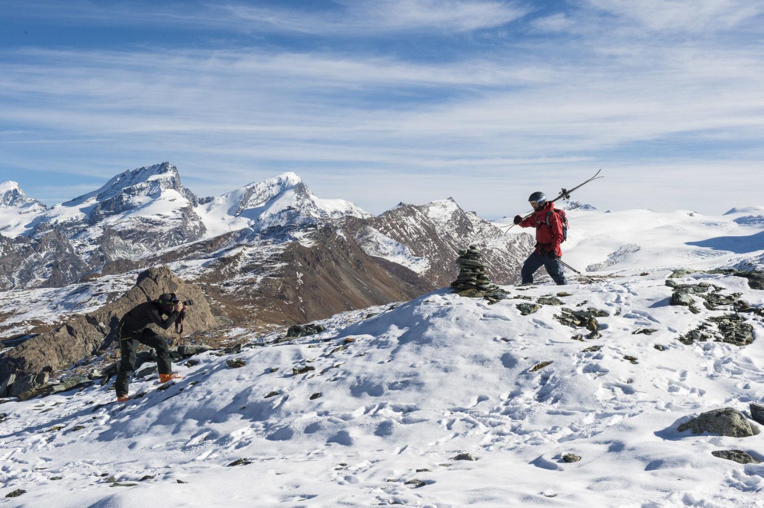 Dos figuras y un perro en la cima de una loma nevada, bajo el sol